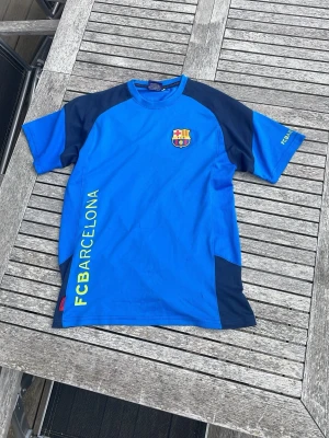 Blå FC Barcelona träningströja - Snygg blå träningströja med mörkblå detaljer och FC Barcelona-logga på bröstet. Trycket 'FC BARCELONA' i gult längs sidan och 'FCB' på ryggen. Perfekt för fotbollsträning eller att representera favoritlaget. Materialet känns lätt och andas bra.