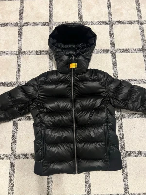 Parajumper jacka - Snygg svart dunjacka från Parajumpers med glansig finish, huva och dragkedja framtill. Jackan har två stora fickor med dragkedja och en cool gul detalj vid halsen. Insidan är ljusgrå och har praktiska innerfickor. Perfekt för kalla vinterdagar.