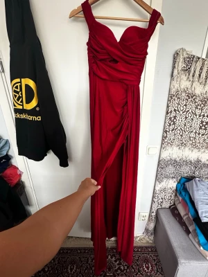 Röd långklänning med slits - Snygg röd långklänning med draperad överdel och breda axelband. Klänningen har en markerad slits framtill och en figurnära passform som framhäver siluetten. Perfekt för dig som vill sticka ut med en elegant look.