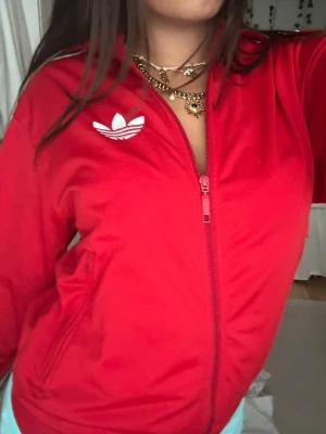 Röd Adidas zip hoodie - Säljer en röd hoodie från Adidas med dragkedja framtill och den klassiska vita trefoil-loggan på bröstet. Hoodien har ribbade muddar och sidofickor. Perfekt för en sportig och avslappnad stil.