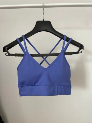 Lila sport-bh med korsade band - Sport-bh i lila med dubbla smala axelband som korsas i ryggen. Tillverkad i stretchigt funktionsmaterial och har bred resår under bysten för extra stöd. Perfekt för träning, yoga eller gym.