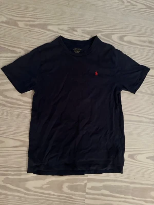 Mörkblå t-shirt från Polo Ralph Lauren - Snygg mörkblå t-shirt från Polo Ralph Lauren med klassisk röd logga broderad på bröstet. Tillverkad i mjuk bomull och har en normal passform med korta ärmar och rund hals. Perfekt till jeans eller shorts för en clean look. Storlek M barn. 150 cm. 