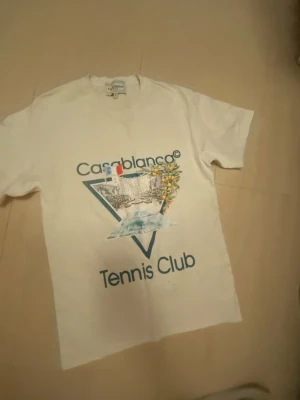 Vit Casablanca Tennis Club t-shirt - Vit t-shirt med tryck från Casablanca Tennis Club. Framsidan har ett stort grafiskt tryck med text, fransk flagga och apelsinblommor. Klassisk passform och rund hals. Perfekt för en sportig och trendig look.