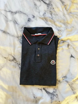 Mörkgrå Moncler piké t-shirt - Snygg mörkgrå piké t-shirt från Moncler med korta ärmar och klassisk krage. T-shirten har vita och röda detaljer på kragen och ärmsluten samt Moncler-logga på bröstet. Perfekt för en stilren och sportig look.