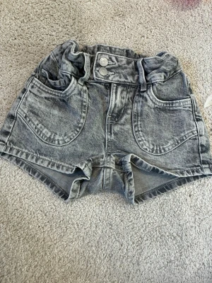 Grå denimshorts med hjärtdetaljer - Säljer ett par grå denimshorts med hög midja och två stora framfickor. Baksidan har två fickor med broderade hjärtdetaljer. Shortsen har dubbel knappstängning och resår i midjan för extra komfort. Perfekta för sommaren!