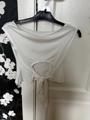Vit croppad topp med knyt och cut-out - Snygg vit croppad topp med cut-out och knyt framtill. Toppen är ärmlös och har en enkel, stilren design som passar perfekt till jeans eller kjol. Tillverkad i mjukt bomullsmaterial för en bekväm känsla.