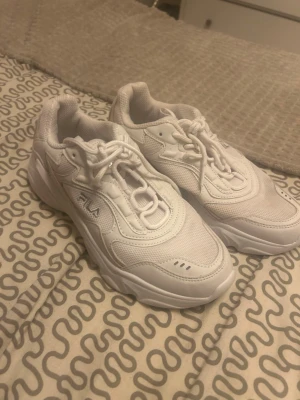 Vita chunky sneakers från FILA - Säljer ett par helvita chunky sneakers från FILA i storlek 40. Skorna har en kraftig sula, mesh- och syntetdetaljer samt klassisk snörning. Perfekta för dig som gillar en sportig och trendig look.