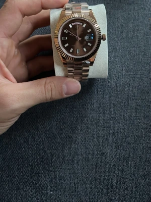 Guldfärgad klocka med länkarmband - Stilren klocka med guldfärgat länkarmband och mörk urtavla. Klockan har tydliga indexmarkeringar, dag- och datumvisning samt räfflad bezel. Perfekt accessoar för dig som vill ha en elegant look.