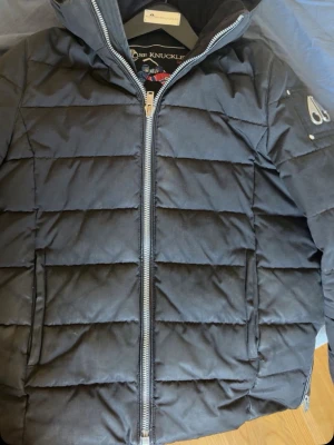 Svart dunjacka från Moose Knuckles - Snygg svart dunjacka från Moose Knuckles med quiltad design och dragkedja framtill. Jackan har en broderad hockeypatch på insidan och en metallogo på ärmen. Perfekt för kalla dagar och riktigt stilren look.