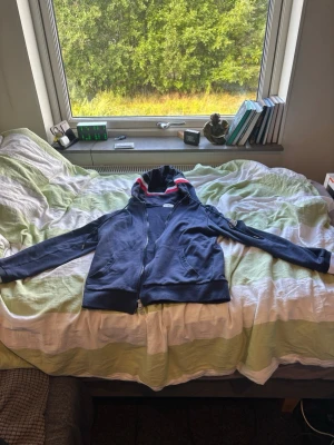 Moncler hoodie med dragkedja - Säljer en Moncler hoodie med dragkedja och huva. Tröjan har röda och vita ränder på huvan och muddar i samma färger. Två fickor framtill och ribbade muddar. Perfekt för en avslappnad och sportig stil.