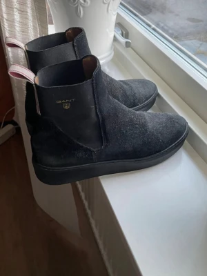 Svarta Chelsea boots från Gant - Snygga svarta Chelsea boots från Gant i mocka med elastiska paneler på sidorna och en diskret logga i guld på utsidan. Skorna har en robust svart sula och en rödvit hälflik för enkel påtagning. Perfekta för en stilren och trendig look. Stl 37 . Perfekt nu när det börjar bli kallare 🍁🍂