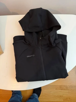 Svart hoodie från Craft med dragkedja - Säljer en svart hoodie från Craft med hel dragkedja och huva. Tröjan har diskret logga på bröstet och ett litet mönster på nederkanten. Materialet känns mjukt och sportigt, perfekt för chill eller träning. Ribbstickade muddar på ärmar och nederkant.