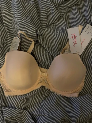 Beige bh från Triumph - Säljer en beige push-up bh från Triumph med vadderade kupor och fina spetsdetaljer längs nederkanten. Bh:n har justerbara axelband och en liten rosett framtill. Perfekt för dig som vill ha både komfort och snygg design.