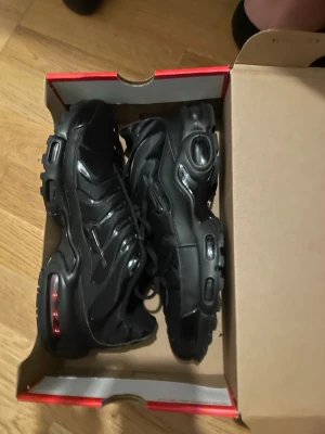 Svarta Nike Air Max Plus sneakers - Säljer ett par helsvarta Nike Air Max Plus sneakers med synliga luftkuddar i sulan och klassisk vågig design på ovandelen. Skorna har en chunky sula och är tillverkade i en mix av syntet och mesh för en sportig look.