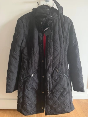 Svart dunjacka från Desigual - Snygg svart dunjacka från Desigual med quiltat mönster, avtagbar huva och dragkedjor vid fickorna. Insidan är röd vilket ger en cool kontrast. Jackan har knappar och dragkedja framtill för extra skydd mot kyla.