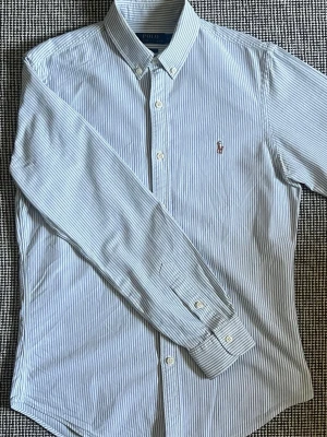 Randig oxford skjorta från Polo Ralph Lauren - Klassisk blå- och vitrandig oxford skjorta från Polo Ralph Lauren med broderad logga på bröstet. Skjortan är i storlek medium och har lång ärm, knappar framtill och button-down krage. Perfekt för en stilren look. Nypris: 1899kr