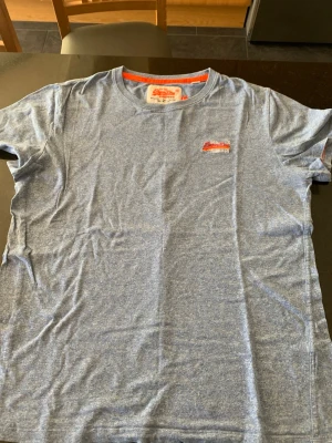 Blåmelerad t-shirt från Superdry - Säljer en blåmelerad t-shirt från Superdry med broderad logga i rött och vitt på bröstet. T-shirten har rund halsringning och korta ärmar. Tillverkad i mjuk bomull, perfekt för en avslappnad stil.