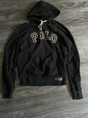 Svart hoodie från Polo Ralph Lauren - Svart hoodie från Polo Ralph Lauren med stor vit POLO-text på bröstet och en klassisk känguruficka. Huvan har vita snören och nederkanten har en liten patch med siffrorna 67. Perfekt för en chill och stilren look.