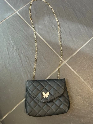 Svart quiltad axelväska med kedja - Snygg svart axelväska i quiltat mönster med guldfärgad kedjerem och en söt fjärilsdetalj på locket. Väskan har magnetlås och är perfekt för att bära det viktigaste. Materialet ser ut att vara konstläder och insidan är enkel och stilren.