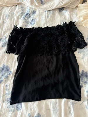 Svart offshoulder spetsblus - Säljer en svart offshoulder blus med blommig spets upptill och transparent tyg nedtill. Blusen har en romantisk vibe med volangdetaljer och är perfekt för dig som vill sticka ut lite extra.