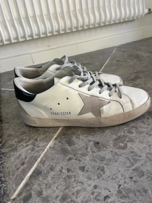 Golden Goose vita sneakers med stjärna - Snygga vita sneakers från Golden Goose med grå mockastjärna på sidan, svarta detaljer på hälen och grå skosnören. Skorna har en klassisk låg profil och är tillverkade i läder och mocka. Perfekta för en trendig och avslappnad look.
