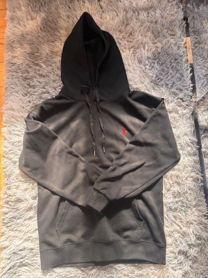 Svart hoodie från Polo Ralph Lauren - Säljer en svart hoodie från Polo Ralph Lauren med klassisk röd logga broderad på bröstet. Hoodien har huva med dragsko, känguruficka och ribbade muddar. Perfekt för en chill och stilren look.