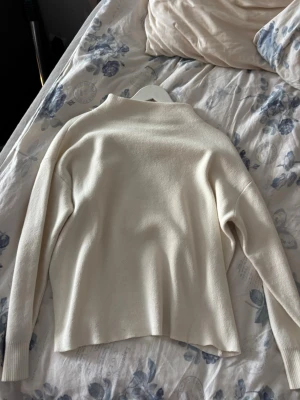 Krämvit stickad tröja med hög krage - En stilren krämvit stickad tröja med högre krage och långa ärmar. Tröjan har en enkel, rak passform och ribbade muddar vid ärmsluten. Perfekt för lager på lager och passar till det mesta i garderoben.