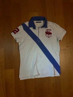 Vit Ralph lauren pikétröja Storlek XS - Snygg vit pikétröja från Ralph Lauren med blå diagonal rand över hela tröjan, blå krage och röd detalj med nummer 2 på ärmen. På bröstet finns ett rött tryck med polospelare och text. Perfekt för en sportig och stilren look.