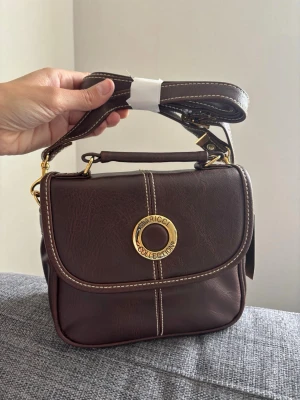 Brun handväska från Nina Ricci - Snygg mörkbrun handväska från Nina Ricci Collection i slätt konstläder. Väskan har guldfärgade detaljer, avtagbar axelrem och dekorativa vita sömmar. Klassisk fyrkantig form med lock och handtag upptill. Perfekt accessoar för en trendig look. Mått 20x18x10 cm 
