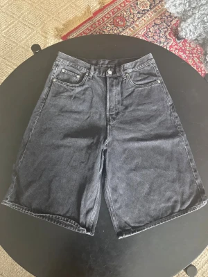Svarta bermudashorts i skate stil - Säljer ett par svarta denimshorts med klassisk femficksdesign och knappgylf. Shortsen har en rak och loose passform med midwaist. Perfekta för sommaren och lätta att matcha med olika stilar. Storlek 30 i Weekday sizing (30-inch waist), ungefär S/M.