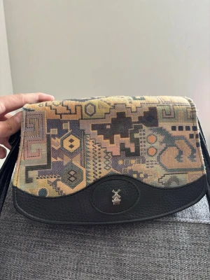 Retro axelremsväska med mönster - Unik handväska med retroinspirerat geometriskt mönster i beige, lila och grönt på locket. Väskan har detaljer i svart konstläder och ett emblem med en kvarn framtill. Axelrem och kropp i grått tygmaterial. Perfekt för dig som gillar vintagevibbar och coola detaljer. Mått 24x16x12 cm