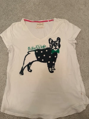Vit t-shirt med hundmotiv Molly & Whyte - Vit t-shirt från Molly & Whyte med ett stort tryck av en fransk bulldogg i svart med vita prickar och en grön fluga. T-shirten har v-ringning och korta ärmar. Perfekt för dig som gillar unika och lekfulla prints.