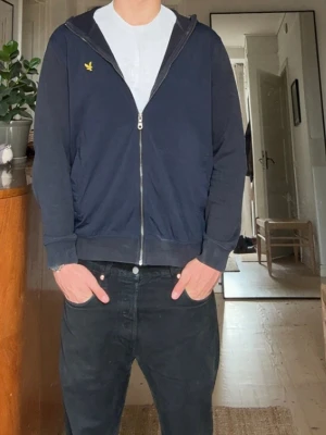 Blå hoodie med dragkedja och huva - Säljer en blå hoodie med dragkedja och huva. Tröjan har ett gult broderat märke på bröstet och ribbade muddar vid ärmslut och nederkant. Perfekt för en avslappnad och stilren look.