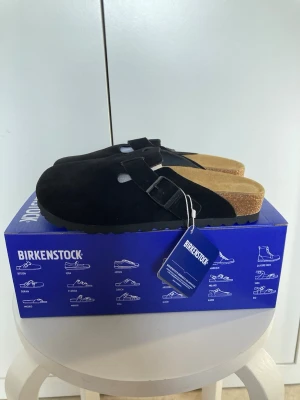 Svarta Birkenstock tofflor mocka - Säljer ett par svarta Birkenstock tofflor i mocka med klassisk slip-in design och justerbart spänne. Fotbädden är i kork och innersulan i mjukt material för extra komfort. Perfekta för dig som gillar stilrena och bekväma skor.