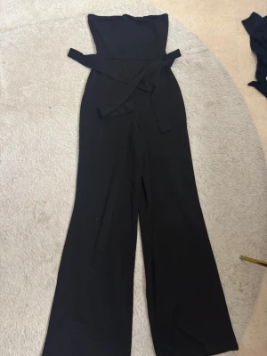 Svart jumpsuit från Nelly  - Snygg svart jumpsuit från Nelly i storlek XS. Modellen har vida ben, markerad midja med knytband och ärmlös överdel med rak halsringning.