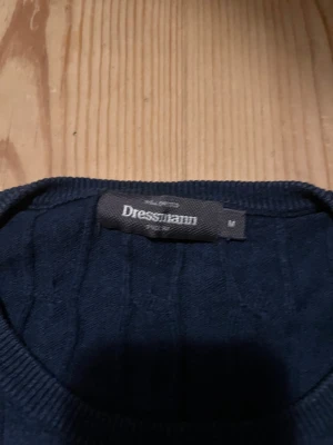 Mörkblå kabelstickad tröja Dressmann - Snygg mörkblå kabelstickad tröja från Dressmann i storlek M. Tröjan har rund halsringning, långa ärmar och ribbade muddar vid ärmslut och nederkant. Perfekt för lager på lager och enkel att matcha med jeans eller chinos.