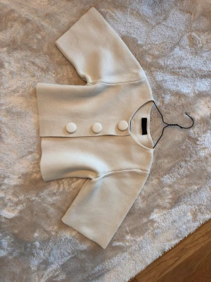 Kort beige kofta med stora knappar - Stilren kort kofta i beige med tre stora tygklädda knappar framtill. Koftan har en boxig passform och vida, halvlånga ärmar. Materialet är tjockt och strukturerat, vilket ger en exklusiv känsla. Perfekt att slänga över en topp för en trendig look.