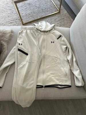Under Armour Tracksuit - Tracksuit i beige från Under Armour. Setet består av en zip-hoodie med fickor och matchande byxor med resår i midjan och svarta dragkedjor. Kan även bytas mot andra plagg/skor.
