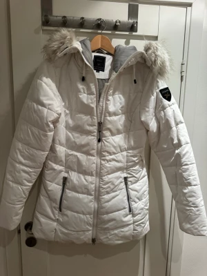 Vit pufferjacka med fuskpäls - Snygg vit pufferjacka med avtagbar huva och fuskpälsdetalj. Jackan har två dragkedjeförsedda fickor framtill och quiltad design. Märke på ärmen och grått innerfoder ger extra stilpoäng. Perfekt för kalla vinterdagar.
