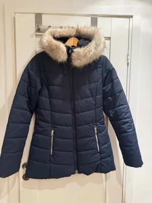 Mörkblå dunjacka med fuskpäls - Snygg mörkblå dunjacka från Canard Collection med avtagbar huva och fuskpäls. Jackan har två dragkedjeförsedda fickor framtill och quiltad design som ger en modern look. Perfekt för kalla vinterdagar och riktigt varm.