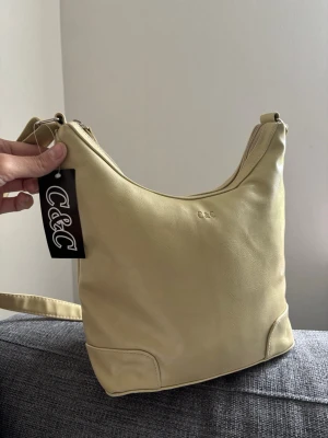 Ljusgul axelväska från C&C - Stilren ljusbeige axelväska från C&C i mjukt konstläder. Väskan har en enkel, rundad form med diskret logga framtill och justerbar axelrem. Perfekt för dig som vill ha en clean och modern look. Stängs med dragkedja upptill. Mått 28x26x11 cm 