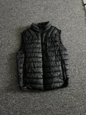 Svart quiltad dunväst - Snygg svart dunväst med quiltat mönster och hög krage. Västen har dragkedja framtill samt två sidofickor och en bröstficka med dragkedja. Perfekt att slänga över en hoodie eller tröja för extra värme. Materialet är glansigt och lätt.