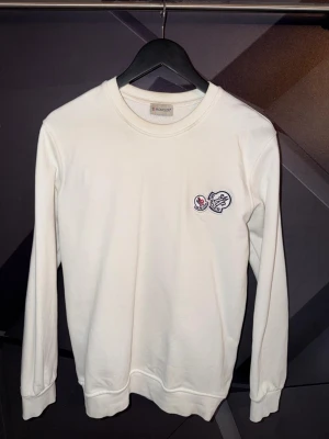 Moncler tröja - Snygg vit långärmad tröja från Moncler med rund halsringning och broderad logga på bröstet. Tröjan har en klassisk passform och är tillverkad i mjukt bomullsmaterial. Perfekt för en stilren och enkel look.