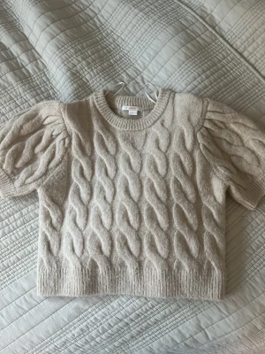 Beige stickad tröja Gina Tricot - Supersöt beige stickad tröja från Gina Tricot med puffärmar och grovt kabelstickat mönster. Tröjan har rund halsringning och ribbade muddar vid ärmslut och nederkant. Perfekt för dig som gillar trendiga detaljer och mjuka material.