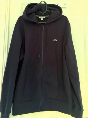 Svart zip hoodie från Lacoste - Säljer en svart hoodie från Lacoste med dragkedja framtill och klassisk huva. Hoodien har det ikoniska krokodilmärket broderat på bröstet och två fickor fram. Tillverkad i mjukt bomullsmaterial och har ribbade muddar vid ärmslut och nederkant.