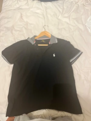 Polo Piké - Snygg svart pikétröja med korta ärmar och vit logga på bröstet. Kragen har grå och vita ränder samt en liten röd detalj.i Perfekt till jeans eller shorts för en clean look.