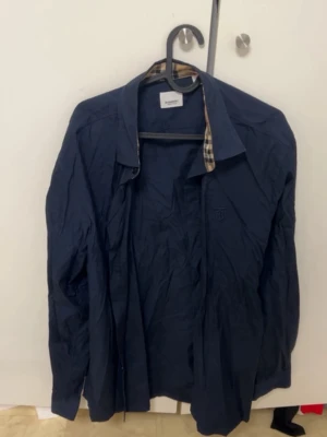 Mörkblå skjorta från Burberry XXL - Snygg mörkblå skjorta från Burberry i storlek XXL. Klassisk Burberry-rutig detalj på insidan av kragen och diskret broderad logga på bröstet. Skjortan har lång ärm och knäppning framtill. Perfekt för dig som gillar stilrena och exklusiva plagg. Behöver strykas och katt hår ska tas bort. 