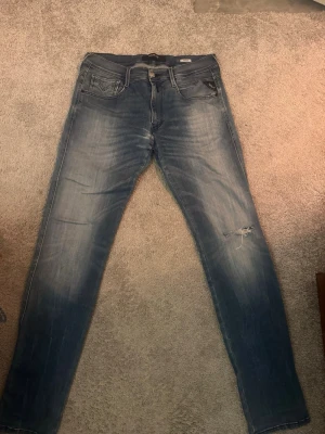 Blå slitna jeans från Replay - Säljer ett par blå jeans från Replay i modellen Anbass. Jeansen har skinny fit, slitningar på höger knä och klassisk femficksdesign. Materialet är denim med stretch för extra komfort. Perfekta för en avslappnad och trendig look. Pris kan diskuteras