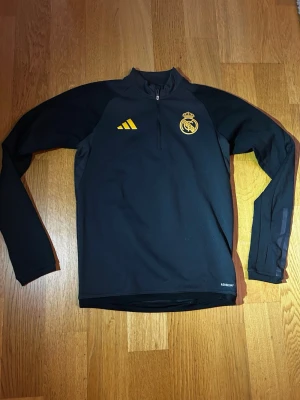 Svart Real Madrid träningströja - Snygg svart långärmad träningströja från Adidas med Real Madrid-logga och gula detaljer. Tröjan har halv dragkedja framtill och är tillverkad i ett tekniskt material som andas. Perfekt för fotbollsträning eller gymmet.