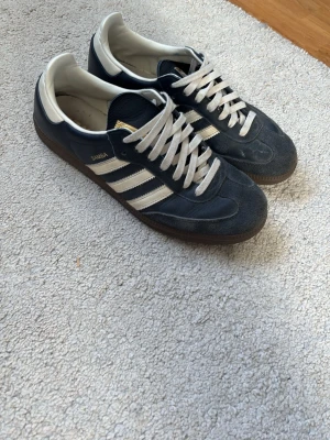 Adidas Samba Blåa sneakers - Klassiska Adidas Samba sneakers i Mörk blå mocka och läder med vita detaljer och de ikoniska tre ränderna på sidan. Skorna har vita snören, gummisula och gulddetaljer vid plösen. Perfekta för en sportig och stilren look.